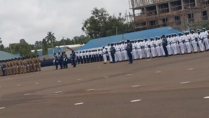 The video shows two groups of military personnel standing in formation on a large asphalt ground. On the left side, the group is dressed in khaki uniforms, while on the right side, they are in white naval uniforms. In the foreground, a man in a black shirt and pants walks toward the camera while holding a DSLR camera in his right hand. In the background, there is a building under construction with scaffolding visible. Other buildings and trees are also present. The sky is overcast and gray. The ground has painted white lines marking the formation areas. Military commands are audible in the background.