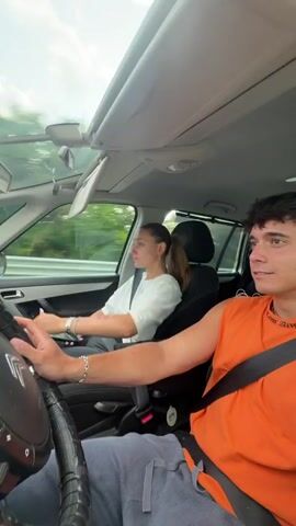 Two people are in a car, a man is driving and a woman is in the passenger seat. The woman is moving her hands and arms in a rhythmic way. The man appears to be looking forward and is not reacting to the woman's movements. The car is moving on a road with trees visible in the background. The sky is overcast.