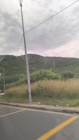 The video shows a view from a vehicle moving along a paved road with yellow lane markings. On the right side of the road, there is a grassy verge and a metal guardrail. Beyond the verge, a lush green hillside covered in trees and vegetation rises. In the background, the sky is overcast with gray clouds. A utility pole with a light fixture is visible on the left side of the road, and a power line stretches across the frame. The camera angle is low, suggesting it is mounted within the vehicle. The movement of the vehicle is evident from the blurred background and the shifting perspective of the scenery.