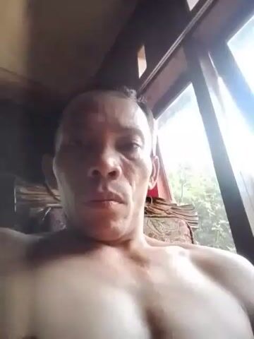 A video shows a close-up of a shirtless man's face and chest. The man is looking directly at the camera. His skin appears sweaty. In the background, there is a window with sunlight streaming through, revealing trees and foliage outside. The man is seated on what appears to be a cushioned chair with a patterned fabric.