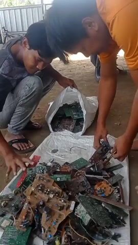 Two men are kneeling and sorting through a pile of electronic circuit boards. The boards are green and brown and some have wires attached. The men are wearing casual clothes. One man is wearing a dark t-shirt and grey pants, the other is wearing an orange t-shirt. They are outdoors in what appears to be a workshop or storage area. There is a white tarp spread on the ground and a white plastic bag filled with circuit boards. In the background, there is a metal structure and a motorcycle wheel.