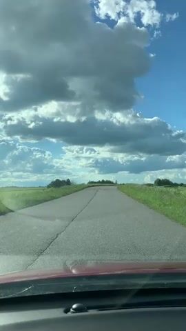 This media file captures a view from a car traveling down a rural road. The subject is the road itself, which appears cracked and worn, stretching towards a distant horizon. Flanking the road are fields of green grass and scattered trees. Above, a dramatic sky dominates the scene, filled with large, billowing white clouds set against a vibrant blue sky. The lighting suggests it is daytime, likely afternoon, with bright sunlight illuminating the landscape and casting shadows within the clouds. The overall mood is one of serene travel through a natural, open landscape. There is no visible text in the media file. The location context provided is Lukaši, Russia, which aligns with the rural, open landscape depicted.