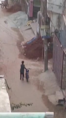 In a high-angle shot, three children are seen interacting with bicycles on a dusty street flanked by buildings and an assortment of debris. One child walks alongside a small, blue bicycle, pushing it forward. Two other children ride bicycles behind him, progressing down the street. Overhead, a network of wires crisscrosses the sky. The area is littered with construction materials, including pipes, tarps, and piles of red dirt. The scene suggests an urban residential area possibly undergoing construction or repair. The light is subdued, casting long shadows on the ground.