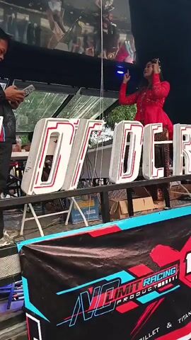 A man in a black jacket with red accents stands on the left, looking at his phone. In the center and to the right, a woman in a red traditional outfit dances, her right arm raised. Large, illuminated white letters spelling "MOTORCRAFT" are displayed on a table in front of them. Behind the letters, a banner with the "NO LIMIT RACING" logo is visible. The scene appears to be outdoors, possibly at an event.
