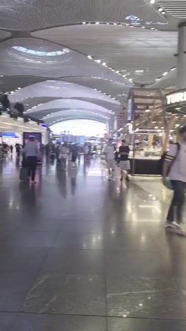 A person walks through a large, modern airport terminal. The terminal features a high, arched ceiling with intricate latticework, and is lit by natural light and overhead artificial lights. Various shops and cafes line the walkway, with signage visible such as "LC Waikiki" and "BLUEMARKET". Several other travelers are present, some walking, some with luggage. The floor is polished and reflective, mirroring the ceiling and lights. The person in focus is wearing a backpack and carrying green shopping bags.