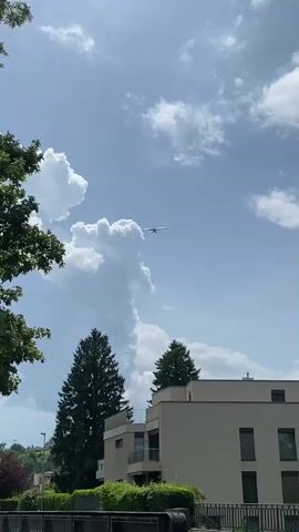 The video shows a small airplane flying in a blue sky with puffy white clouds. The plane appears to be approaching the viewer. In the foreground, there is a leafy green tree, a modern building, and a signpost with directions to several locations including "Embouchure de l'Areuse" and "Plage de Boudry". The sky is bright and sunny.