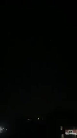 The video shows a dark night scene with a few faint lights on the horizon. There is a building in the foreground to the right. Suddenly, the screen fills with bright white light.