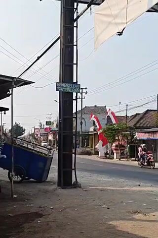 The video shows a street scene with a utility pole in the foreground. A blue cart is on the left side of the road. There are buildings and shops on the right side of the road, decorated with Indonesian flags. Several motorcycles are visible on the road, with people riding them. The sky is clear and the sun is shining. A sign on the utility pole reads "BENGKEL LAS TAMBRAN".