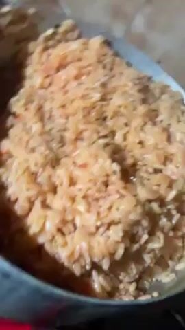The camera angle is from slightly above, showing a pot of cooked rice being stirred on a stovetop. The rice appears reddish-orange and moist. The pot is metallic, with signs of use around the rim. The stovetop underneath the pot is black. The background is blurred, but appears to be a wall with a patterned surface, possibly tile. The stirring motion is subtle and does not reveal any identifiable people.