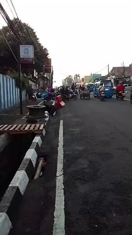 The camera pans across a busy street in what appears to be an urban or suburban area. The initial view shows a sidewalk with black and white striped curbing, a partially covered drainage ditch, and a pallet on the edge. The street is filled with traffic, including cars, vans, and motorcycles. There are people on the sidewalk and on the street. The camera pans to the right, following the flow of traffic and revealing more of the street, adjacent buildings, trees, and billboards. The overall atmosphere seems busy and crowded.