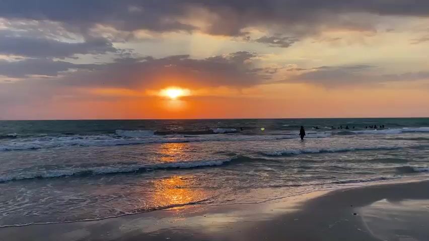A wide shot captures a serene beach scene at sunset. The sky is painted with vibrant hues of orange and pink as the sun, partially obscured by clouds, dips towards the horizon. Rays of light pierce through the clouds, casting a warm glow on the water. Gentle waves lap at the sandy shore, creating a peaceful ambiance. In the distance, a few individuals are visible in the ocean, enjoying the water. One person, standing in the shallows, raises their arms in a gesture of triumph or joy. The reflection of the setting sun shimmers on the wet sand in the foreground, adding to the picturesque view.