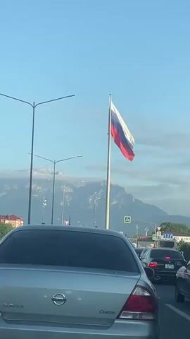 The video shows a view from inside a vehicle, likely in traffic. In the foreground, the back of a silver sedan is visible, with its taillights illuminated. To the right, other vehicles are in traffic, including a black sedan and what appears to be a truck. A Russian flag is prominently displayed on a flagpole to the right of center. In the background, there are streetlights and a mountain range with clouds partially obscuring its peaks. The sky is a clear blue. There are no visible people. The scene suggests a roadside view with a national flag and natural scenery.