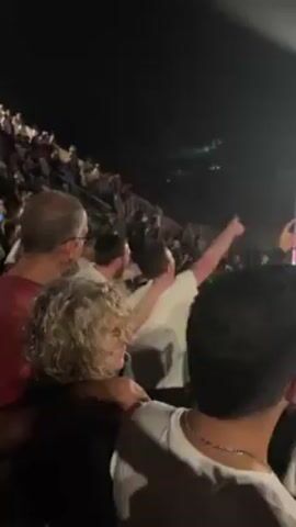 The video shows a crowd of people at an outdoor concert. It is night. The camera is positioned behind a group of four people, looking towards the stage where a performer is visible in the distance. The crowd is dense and many people are cheering. A man in a white shirt is raising his fist in the air. A woman with curly hair is looking towards the stage. An older man with glasses and a red shirt is partially visible on the left side of the frame. The camera is mostly still.