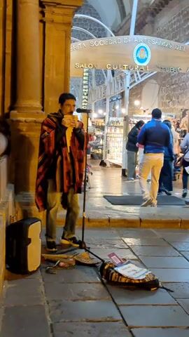 The video shows a street musician playing a pan flute in what appears to be a historic city center. The musician, a man with dark hair, stands on a stone sidewalk near a building with a stone column. He's wearing a colorful poncho, jeans, and sneakers, and is playing into a microphone attached to a stand. To his left is a small speaker. On the ground is a collection of other pan flutes and a bag with a sign on it, likely for donations. Behind the musician is an archway leading into an arcade of shops, with people browsing the stalls. The sign above the archway is partially visible, reading "ERITA SOCIEDAD DE ARTESANOS DEL SALON CULTURAL." The lighting suggests it is evening or a dimly lit area. Several people are seen walking past in the background.
