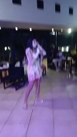 A young woman with long dark hair stands in the center of the frame, holding a microphone and singing. She is wearing a short, brightly colored dress with a floral pattern and silver sandals. The scene appears to be an indoor gathering, possibly a party or a restaurant, with other people visible in the background. Some people are seated at tables, and others are standing. The lighting is dim, with overhead lights illuminating the area. The floor is tiled, and there are chairs and tables scattered around the room. A fan is visible on the ceiling.