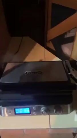 Here is a description of the video:

The video shows a DeLonghi grill being opened to reveal two pieces of meat cooking on the grill's surface. The grill has a digital display showing temperature and settings, as well as knobs for controlling the cooking process. Steam rises from the meat as it cooks, and the grill makes beeping sounds.