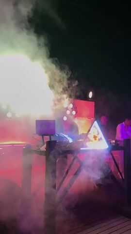 The video opens with a shot of a DJ set up on a raised platform. The DJ is wearing a blue shirt and a baseball cap. They are standing behind a laptop and a DJ mixer, with smoke and pink lights creating a dramatic atmosphere. There are circular lights on the wall behind the DJ. The next shot is a close-up of a person holding a drink, their face obscured by smoke and the bright lights from the stage. The final shot is of the back of a person's head, wearing a plaid shirt and a baseball cap, as they walk away from the camera. The scene appears to be a live music event or club setting with a focus on vibrant lighting and a lively atmosphere.