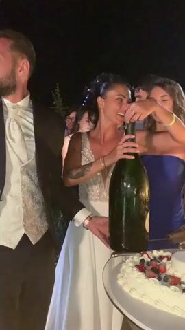 A man in a suit and a woman in a white dress are standing next to a cake and a large bottle of champagne. The woman is attempting to open the bottle of champagne. There are other people in the background, some of whom are holding up phones, presumably to take pictures. The scene appears to be outdoors at night, possibly at a wedding reception, given the presence of a wedding cake and formal attire.