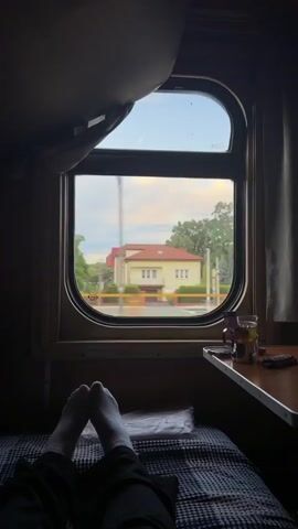 The video shows a view from inside a train compartment. The perspective is from someone lying down on a bunk bed, with their feet, clad in white socks, visible in the foreground. The bed has a dark and white patterned blanket. To the right, a small wooden table is attached to the wall, with a glass of amber liquid and a small dark object on it. The window of the train compartment is oval-shaped and has a thick frame. Through the window, a landscape is visible, consisting of houses with red and yellow roofs, green trees, and utility poles. The scene outside appears to be moving from right to left, indicating the train is in motion. The lighting suggests it is either early morning or late afternoon. A dark curtain or shade is partially pulled down over the top of the window.