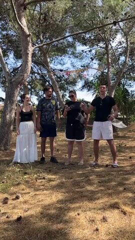 Four people, three males and one female, are standing on a patch of brown ground covered in pine needles and cones beneath a pine tree. The woman on the left is wearing a black tank top and a long white skirt. Next to her is a man in a gray patterned t-shirt and dark blue shorts. The woman in the middle is wearing a black t-shirt and black shorts. The man on the right is in a black t-shirt and white shorts, holding a stick decorated with red and pink paper. In the background is a hammock and other trees. The man on the right is pointing at the camera while the others look towards him. He then makes a "peace" sign with his fingers.