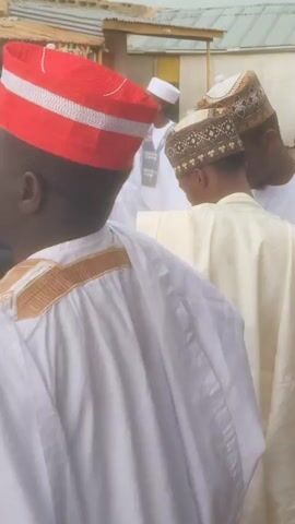The video captures a scene featuring several men dressed in traditional attire, likely in Jogana, Nigeria. The camera mainly focuses on the backs and sides of the men as they move or gather outdoors on a dirt path.

The subjects are adult men, predominantly wearing long, loose-fitting robes or kaftans in white or light cream colors. Their headwear is particularly striking:
*   In the foreground, a man wears a vibrant red cap with broad white horizontal stripes. His white robe features gold embroidery on the shoulders.
*   Immediately behind him, another man wears a dark cap intricately patterned with geometric designs in shades of brown, black, and possibly gold.
*   Further back, a man wears a simpler, plain red cap.
*   Another man is seen in a white and light-striped shirt, also with a red cap.

The setting appears to be a village or small town, characterized by simple, single-story buildings with corrugated iron roofs visible in the background. The ground is unpaved, suggesting a rural or semi-urban environment. The activity seems to be a social gathering or a procession, as the men are walking slowly, congregating, and there is ambient chatter.

The time of day is clearly daytime, with bright but diffuse natural light indicating it could be mid-morning or afternoon. The weather appears clear. A faint, partially obscured sign with the letters "SPA" on a green background is briefly visible on the left side of the frame. The overall atmosphere suggests a community event or daily life in Jogana, Nigeria.