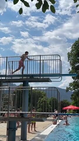 A person in pink swimming trunks jumps from a diving board at a swimming pool. The person performs a flip in the air before splashing into the water. Other people are swimming and relaxing in the pool and on the deck. There are trees and mountains in the background. The sky is blue with white clouds.
