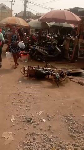 The video shows a crowded marketplace in what appears to be an African country. The ground is dirt and scattered with debris. In the foreground, there is a damaged motorcycle lying on its side. Several people are walking around, including a young woman carrying a white bucket. She is wearing a floral dress and sandals. Another person is carrying a pair of sandals. There are many motorcycles parked in the background. Several vendors are selling goods from under umbrellas, including a cart full of lemons.