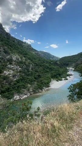 The video shows a river flowing through a mountainous landscape under a partly cloudy sky. The river is a clear turquoise color and flows over a rocky riverbed. The mountains are covered in lush green vegetation, with rocky cliffs and outcrops visible. Two people, a man and a woman, are walking across a narrow, somewhat dilapidated suspension bridge that spans a gap in the rocky terrain. The woman is wearing a red shirt and black shorts, while the man is wearing a light blue shirt and dark shorts. They appear to be walking carefully, holding onto the sides of the bridge for balance.