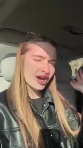 A woman is recording a selfie video inside a car. She is wearing a black jacket and has long, light brown hair and red nail polish. Another woman with brown hair and sunglasses is visible in the background. The woman in the foreground is singing along to the music playing in the car and gesturing with her hands. The car seems to be moving. The environment outside the car is visible through the windows.
