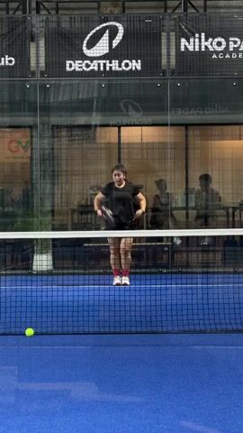 A woman in a black t-shirt and shorts, holding a padel racket, stands on a blue padel court. She is celebrating, with her arms raised and a wide smile on her face. Behind her, a net divides the court, and glass walls enclose the playing area. The background shows a sports facility with branding visible, including "DECATHLON" and "NIKO PADEL ACADEMY." A yellow padel ball rests on the court in the foreground. The woman's expression and posture indicate excitement or victory.