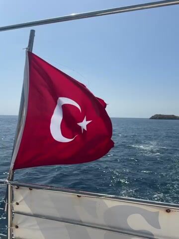 The video shows a boat moving on the sea. A Turkish flag waves in the breeze. In the distance, rocky islands and a coastline covered in greenery are visible. The sky is a clear blue. The wake of the boat is visible in the water.