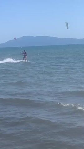 The video shows a person kitesurfing on a blue body of water. The kitesurfer is wearing a dark top and light-colored shorts, and is on a board, generating a spray of water. A kite is visible above the person, connected by lines. In the distance, there are mountains and the sky is clear and blue. There are other kites and windsurfers visible further out on the water. The waves are small, and the water appears calm. The overall scene is a sunny day at the beach, with various watersports activities taking place.