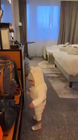 The video shows a small child in a white hooded jumpsuit standing next to an orange-trimmed cabinet in a hotel room. The cabinet contains a backpack and other items. The child is looking at the cabinet and appears to grab something from it. The camera pans across the room, showing a TV, a small round table, a gray padded stool, and two beds with white linens. Each bed has folded white robes on top. The room has gray carpeting, light-colored walls, and curtains covering the window. The lighting is soft, with LED strip lighting along the ceiling.