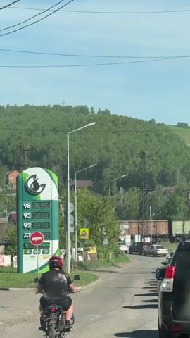 The video captures a daytime scene in Zykova, Russia, under a clear blue sky. A person wearing a red helmet and dark clothing is seen riding a motorcycle away from the camera on a paved road. To the left, a prominent green and white gas station sign displays fuel prices: 98 at 86.90, 95 at 68.90, 92 at 64.90, and AT (likely diesel) at 0.00. Below the prices, a red "no entry" sign with a white horizontal bar is clearly visible, next to a yellow triangular warning sign. On the right, several cars are present on the road, with a dark-colored SUV partially visible in the immediate foreground. In the background, a large, tree-covered green hill rises, dotted with houses. Utility poles and power lines crisscross the frame. Further back, train tracks run parallel to the road, with several freight train cars parked on them.
