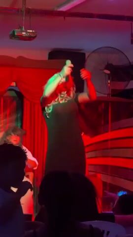A drag performer with blonde hair and heavy makeup is on a stage under colorful lighting. They are wearing a black dress and holding their hands to their head. In the background, there are red curtains and a striped backdrop. Other performers or audience members are visible in the foreground and to the left, mostly silhouetted. The stage appears to be indoors in a performance venue.