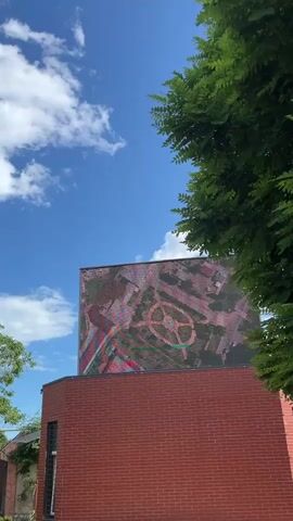 A digital billboard displays an aerial view of a circular garden with pathways and a central feature. The billboard is mounted on a red brick building, partially obscured by the branches and green leaves of a tree in the foreground. The sky above is blue with scattered white clouds. The video captures a still shot of the billboard and its surroundings.