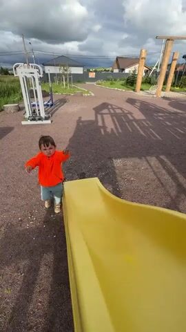 A child in an orange jacket and blue jeans plays on a playground with a yellow slide. The child is picking up small stones and throwing them. The playground has a gravel surface and is surrounded by grass and trees. The sky is cloudy.