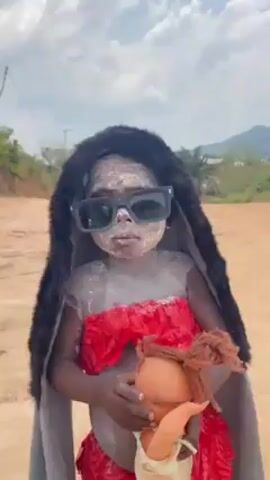 The video features a small child dressed in costume standing on a dirt road in a rural environment. The child's face and body are covered in white powder, and they are wearing oversized black sunglasses and a black furry hood. They are also wearing a red outfit with ruffles and a long grey cape. The child is holding a doll in their arms. The dirt road is surrounded by trees and vegetation, and a mountain is visible in the background under a partly cloudy sky.  The camera zooms out slowly from the child.