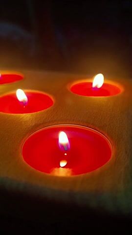The video shows six red tea candles burning in a light wooden holder. The candles are arranged in two rows of three. The flames flicker, and the red wax is visible in each cup. The scene is dark, lit only by the candlelight. Sounds of music are heard throughout the clip.