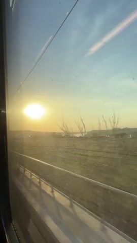 The video shows a view from a train window. The sun is setting and casting a bright glow across the sky. The landscape outside is green and appears to be farmland. A railing runs along the bottom of the window, and the train tracks are visible below. The sun is partially obscured by a distant hill or tree line, and its light creates a lens flare on the window. The sky is a gradient of blue to orange, with some clouds visible. The overall impression is one of a peaceful and scenic journey.