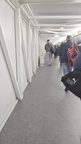 The video captures a group of people walking through an enclosed walkway with white walls and a grey floor. The walkway is illuminated by overhead lights, and its structure is supported by white triangular beams along the walls. The individuals are walking in the same direction, away from the camera. They are dressed in casual clothing, and some are carrying backpacks. A white object that may be an air conditioner or heater is situated against the wall on the left side of the walkway. The overall impression is of a busy thoroughfare, likely in a transit hub or similar facility.
