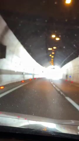 The video shows a first-person perspective of driving through a concrete tunnel. The tunnel is illuminated by overhead lights, and the road ahead is bright as the vehicle approaches the exit. There are red and blue lights along the sides of the road within the tunnel, presumably for traffic guidance. As the vehicle exits the tunnel, the bright daylight reveals a highway with cars and a mountainous landscape in the distance. A road sign indicating a directional arrow is visible on the right side of the highway.