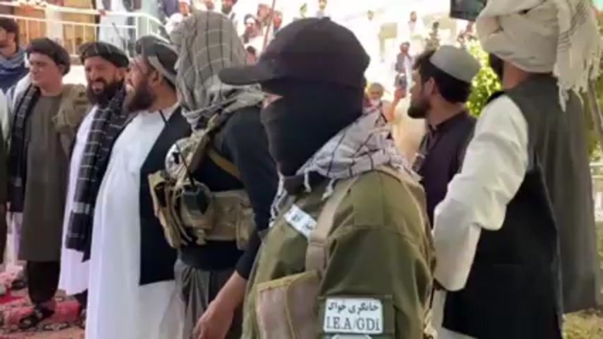 Here's a detailed description of the video:

**Scene:** The video appears to depict a gathering of people in an outdoor setting. A white canopy or awning provides partial shade.

**People:** The video features several men, many of whom seem to be part of a formal group or gathering. Several of them wear traditional Afghan clothing, including long shirts, vests, and turbans. One man is wearing a black baseball cap with a repeating Louis Vuitton symbol pattern, a tactical vest and a green uniform jacket. He also has a face mask covering the lower part of his face. A patch on his uniform is visible, with text written in a non-English script, possibly Pashto or Dari, and the abbreviation "L.E.A./GDI." Some individuals are taking photos or videos with their mobile phones.

**Objects:** Visible objects include traditional Afghan clothing, mobile phones, and military-style vests with pouches.

**Actions and Interactions:** The men seem to be standing and observing something, possibly a speaker or event. The overall atmosphere appears calm and observational.

**Environment:** The environment is bright and sunny. The architecture in the background suggests a building or structure with a covered area.