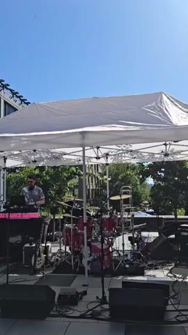 This video begins with a view of a portable canopy set up outdoors, likely for a performance or event. Underneath the canopy, a DJ setup with turntables and a laptop is visible. To the right of the DJ, a drum set and several musical instruments, including guitars and keyboards, are arranged on stage. The area appears to be an outdoor plaza with tiled flooring. In the second part of the video, the camera pans to show a young woman wearing sunglasses and a baseball cap. She is holding a smartphone in her left hand and looking up and to her right. The background shows an urban environment with buildings and trees.