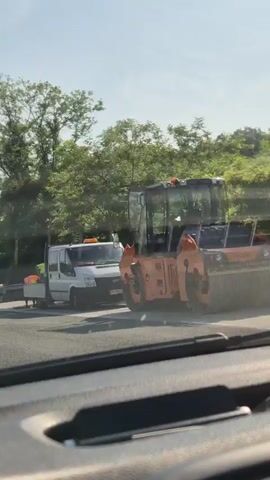 The video shows a construction scene on a sunny day. In the foreground, we see the interior of a vehicle, likely a car, from the perspective of the driver. Orange traffic cones are visible on the road. Further ahead, a white truck with an orange canopy is parked, and two individuals wearing high-visibility vests are near it, seemingly engaged in work. To the left, a piece of construction equipment, possibly a bobcat, is present, and another person in a high-visibility vest is operating it. The road appears to be under repair or construction, with various traffic signs and barriers in place. Trees and foliage line the background, indicating an outdoor, possibly rural or suburban, location. The sky is clear and bright. The overall scene depicts road maintenance or construction activities.