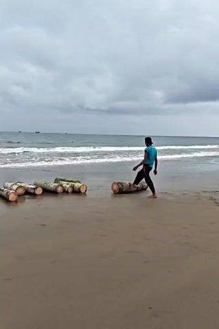 A person in a blue shirt and dark pants walks along a wet sandy beach towards a pile of several logs. The person bends down and grips one of the logs. The person then lifts and rolls the log to the left. The ocean is in the background with waves breaking on the shore. The sky is cloudy.