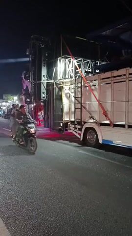 A nighttime street scene features a large truck equipped with a massive sound system and lighting rig. The truck is parked on the right side of the road. On the left side of the road, a person in a white shirt stands near some lights and equipment. Several motorcycles are present on the road, some with riders and passengers. One motorcycle in the foreground has its rear visible, with luggage strapped to it. The lighting is predominantly purple and white, illuminating the sound equipment and the surrounding area. The road surface appears to be asphalt.