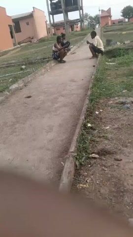 The video shows a low-angle view of a narrow, dirt pathway with concrete borders, bordered by grassy areas and buildings, including a water tower. Three people are squatting along the pathway. One individual wearing a white top and patterned shorts is sitting on the left side, with another person in a dark outfit sitting behind them. A third person in a yellow shirt and dark shorts is squatting on the opposite side, facing the other two. The person in the yellow shirt appears to be talking and gesturing. The person in the white top laughs in response to the person in the yellow shirt.