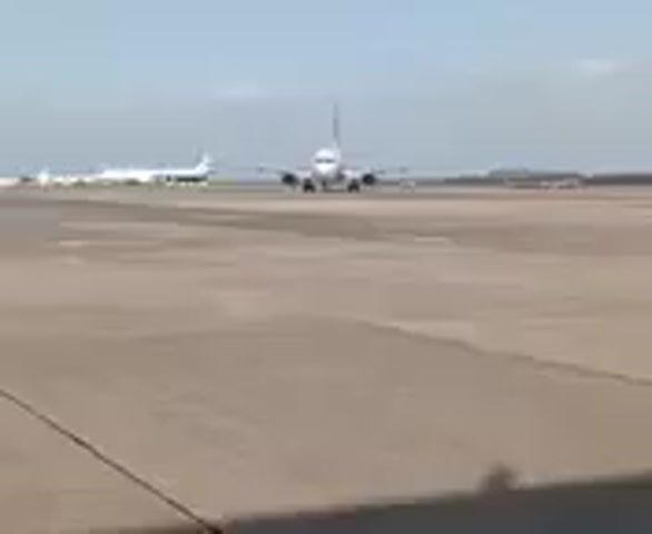 The video shows a passenger jet moving slowly on a concrete runway. The plane is white with dark markings. There are other aircraft visible in the background, also on the runway. The sky is light blue with some thin clouds. The camera is stable, and the scene is viewed from a low angle.