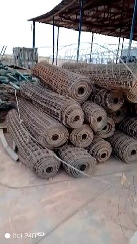 The video shows a fenced outdoor industrial area containing construction materials and equipment. Initially, a large pile of rolled wire mesh lies on the ground beneath a covered structure. The camera then pans to show a pile of metal bars and wire. A small gasoline-powered engine sits on a wooden pallet near a concrete mixer. Finally, the camera shows the concrete mixer located on a metal sheet near debris.